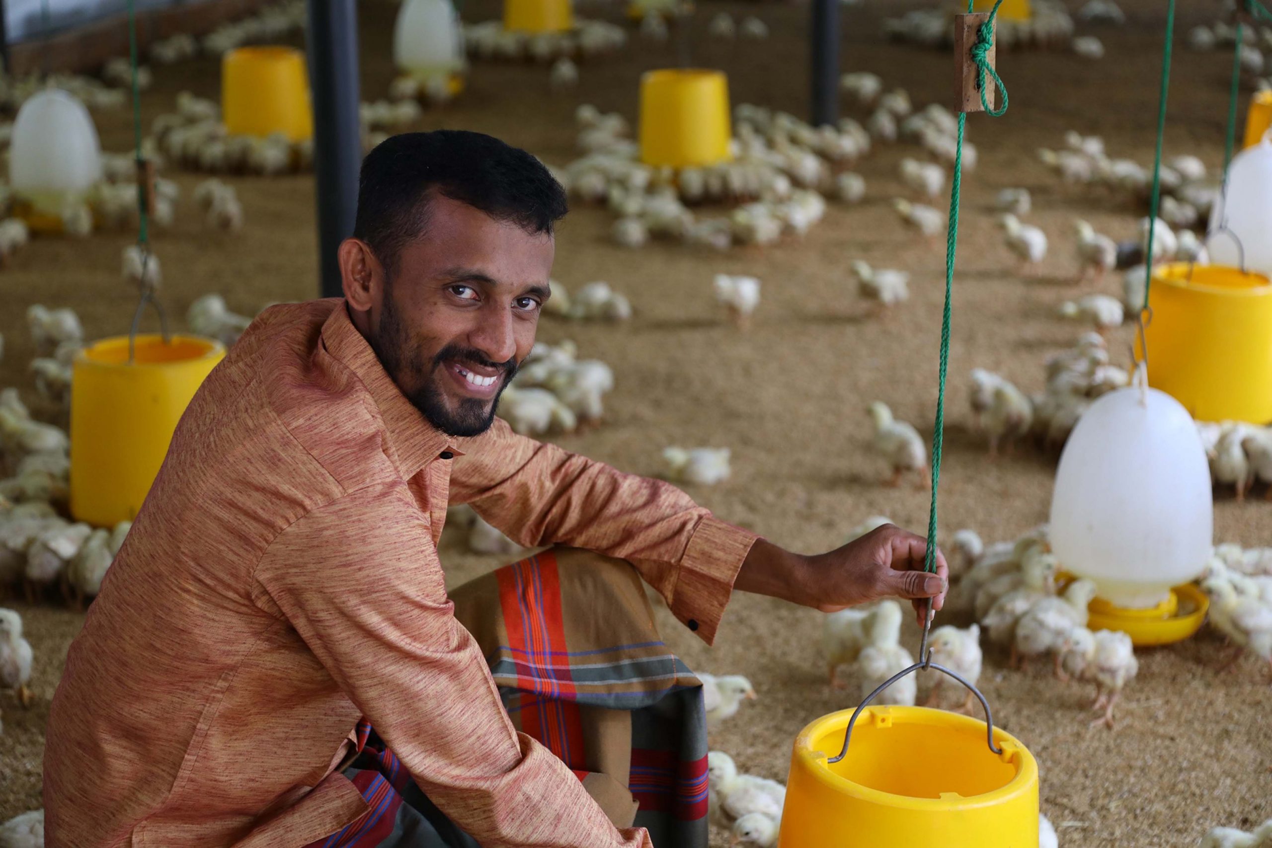 Man sitting in a baby chicken area smiling at the camera while chicks feed from feeders around him.