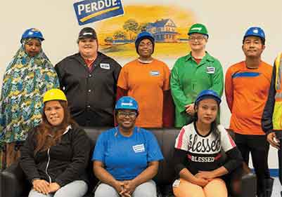 Group shot of eight people in hard hats stand and sitting in front of a wall with the Perdue Agribusiness logo on the wall - showcasing diversity.