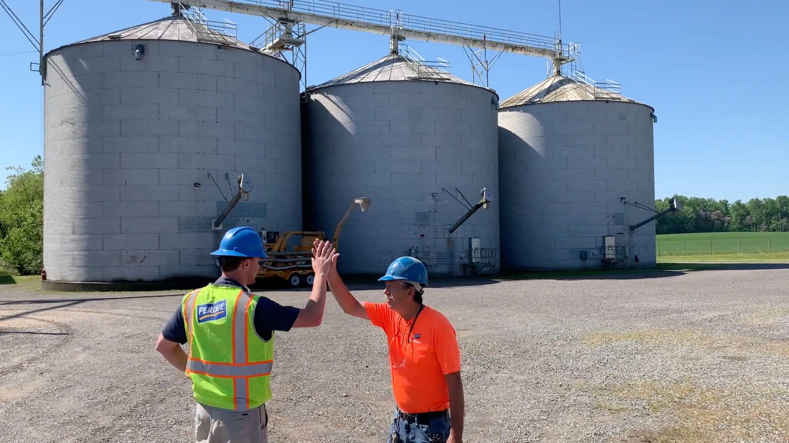 Man in yellow reflective Perdue vest and man in orange t-shirt with blue helmets high fiving in front of 3 industrial silos.