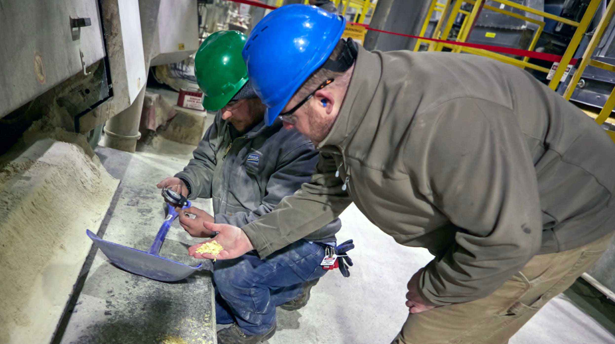Two men in hardhats in a factory setting. One kneeling by a shovel looking at a gauge and one leaning over looking at material in the palm of his hand.