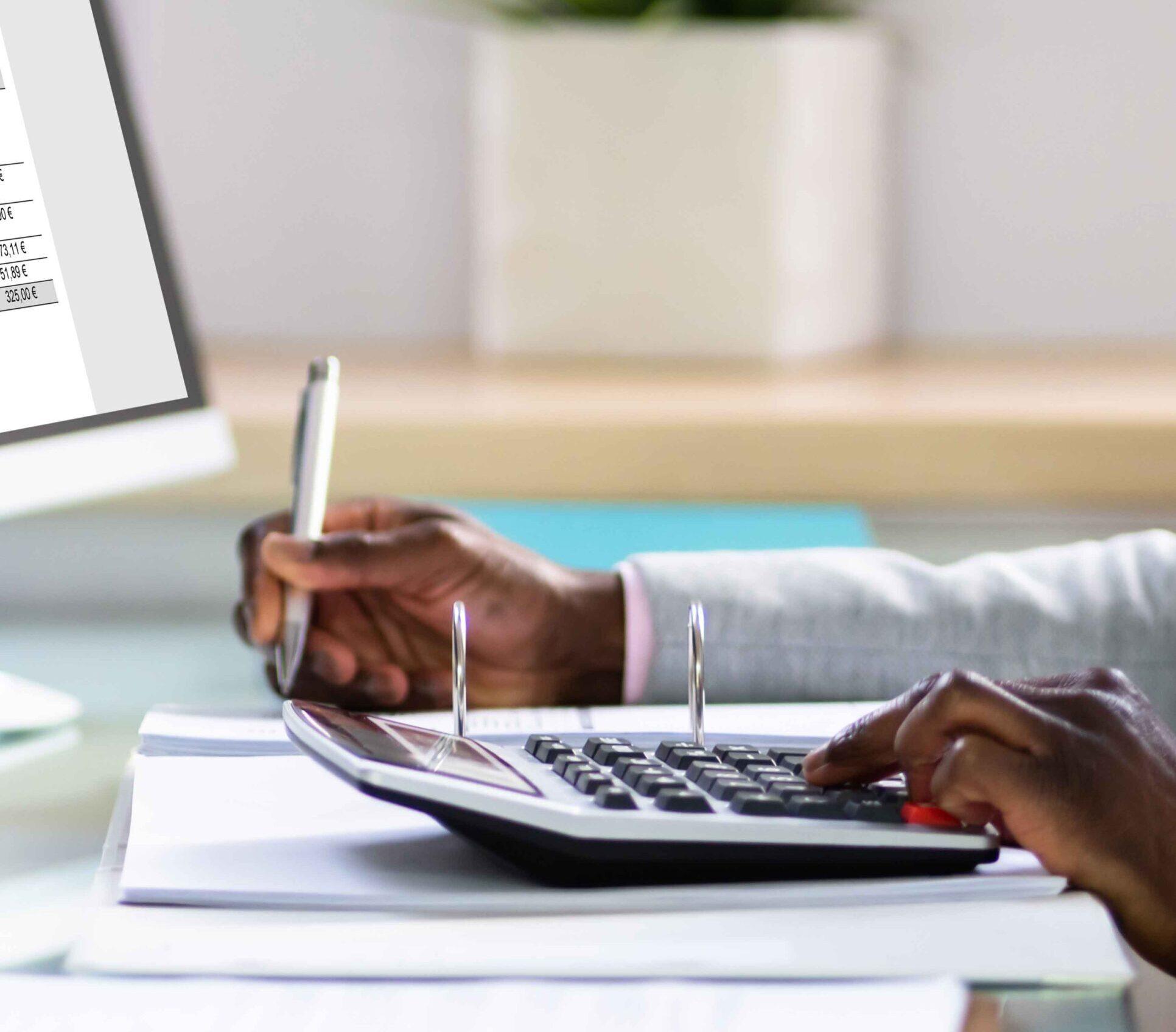 Desktop with computer and papers with one hand holding a pen and another hand on a calculator on it